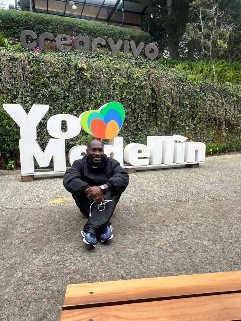Man smiling while seated in front of the “Yo Medellín” sign at Parque Arví, surrounded by lush greenery.